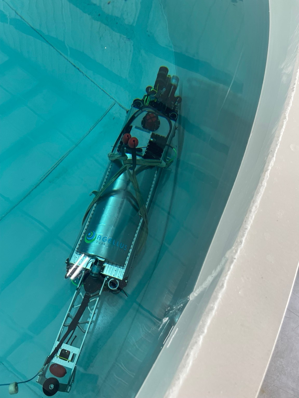 Agellus tank-inspection robot submerged beside a curved pool wall, with its long frame and cylindrical housing visible.