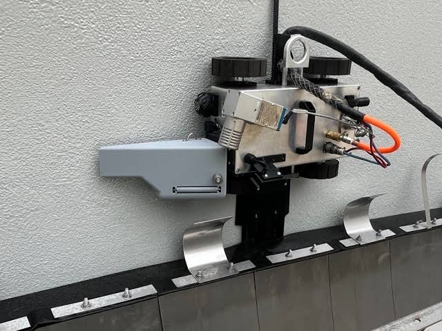 Pike Robotics inspection robot against a textured vertical wall, with a metal housing, gray side-mounted enclosure, and visible cabling.